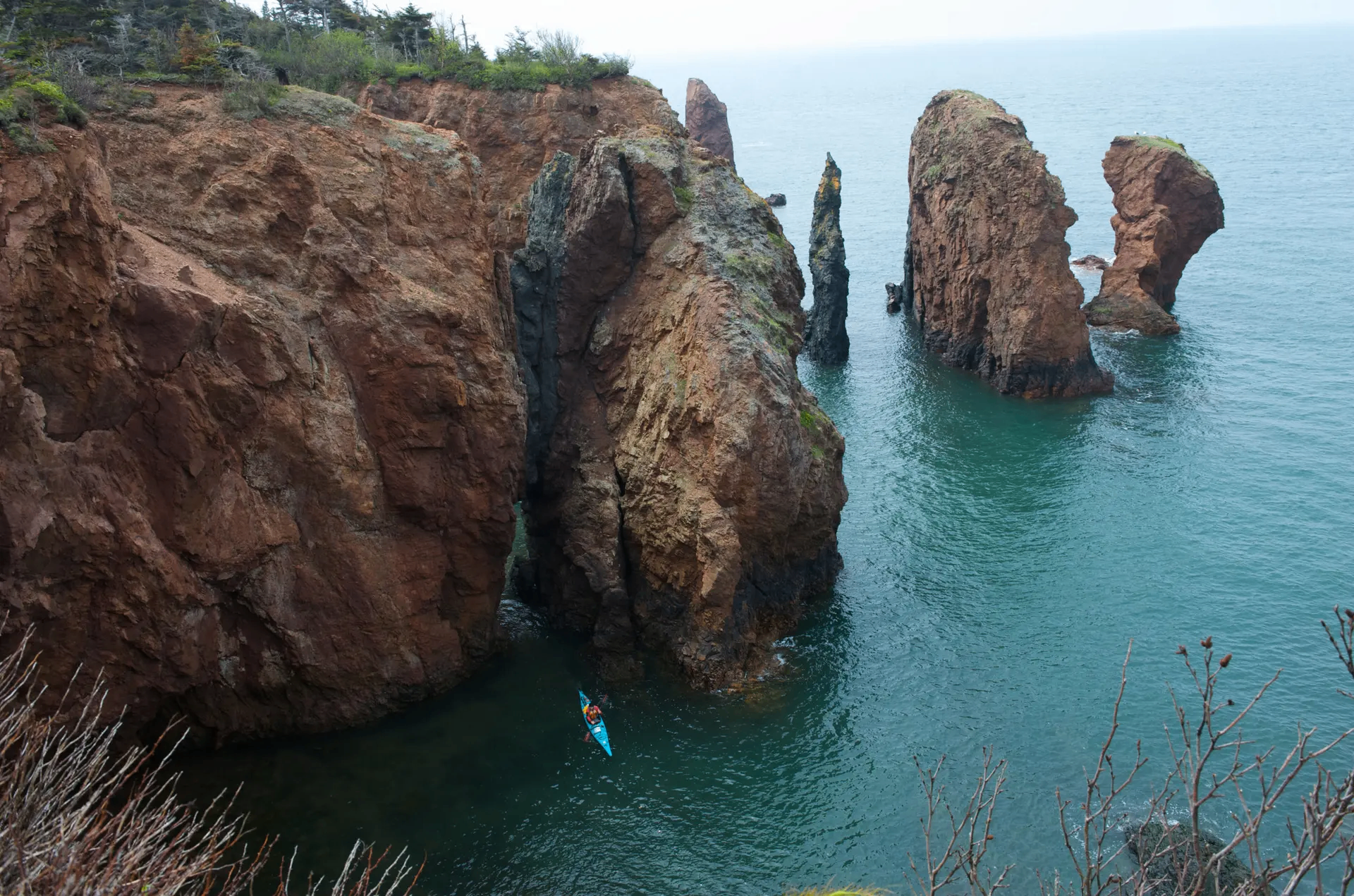 Aerial view of a sea kayaker paddling below towering red sandstone sea stacks and cliffs on the Bay of Fundy