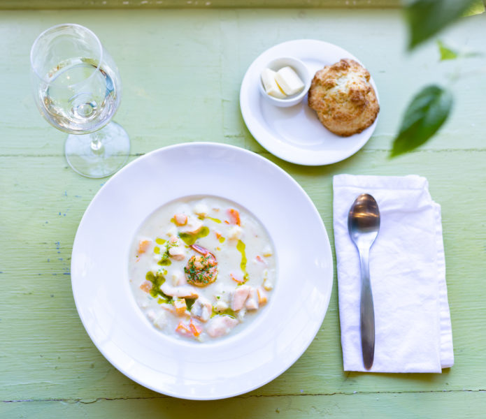 Bowl of seafood chowder with shrimp and salmon, biscuit, butter and a glass of white wine on a green table